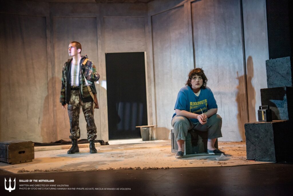 A scene from the play Ballad of the Motherland. A soldier stands to the left in a prison space. He is watching over a young prisoner, a Canadian girl named Kate, who sits on a wooden box to the right. Neither look at the other. Kate looks directly at the audience. The soldier looks away to the left. 