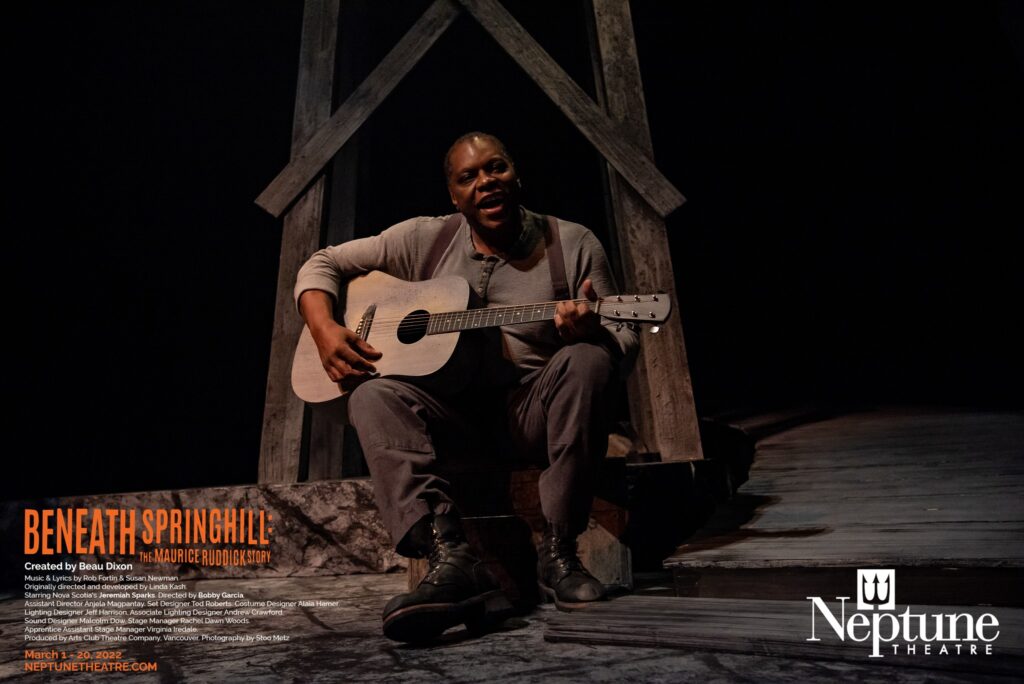 A production photo from Beneath Springhill at Neptune Theatre. A black man sits in a mine shaft playing the guitar. He is dressed in 1940s style brown muted clothing. He smiles as he sings. 