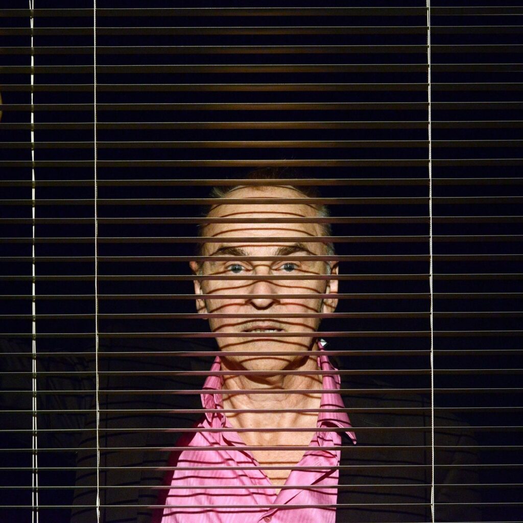Daniel MacIvor, a white cis actor in his 50s, stands behind a venetian blind. The slats are open so we can see his face- especially prominent eyebrows and intense eyes. He is wearing a bright pink button down shirt. 