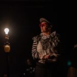 A non-binary actor in a cap with red glasses and a Elizabethan Clown-adjacent shirt sits to the right of a ghost lamp on a darkened stage.