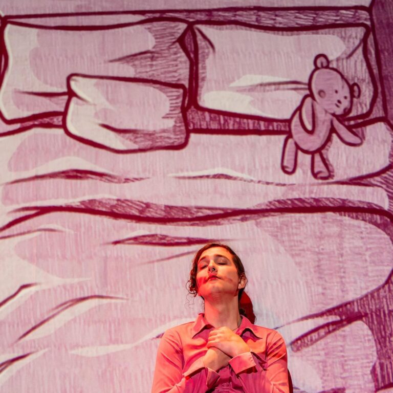 A trans woman in a pink blouse stands in front of a projected cartoon bed with a teddy bear on it. The trans woman clutches her hands over her heart.