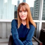 A white woman sits with her arms folded in front of her looking at the camera. She has reddish blonde hair with bangs, and an intense expression. She wears a blue sweater which sets off the colour of her hair beautifully.