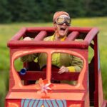 A woman in a green outfit with huge old fashioned riding goggles on sits exuberantly inside a big cardboard car. She is playing Toad in Wind in the Willows.