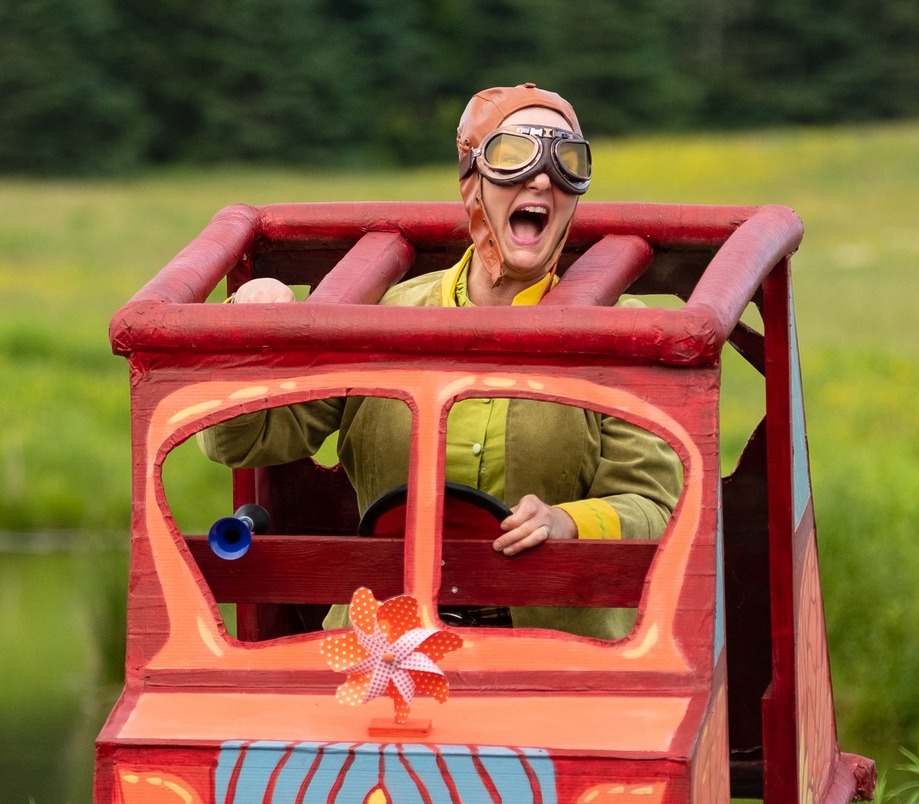 A woman in a green outfit with huge old fashioned riding goggles on sits exuberantly inside a big cardboard car. She is playing Toad in Wind in the Willows.