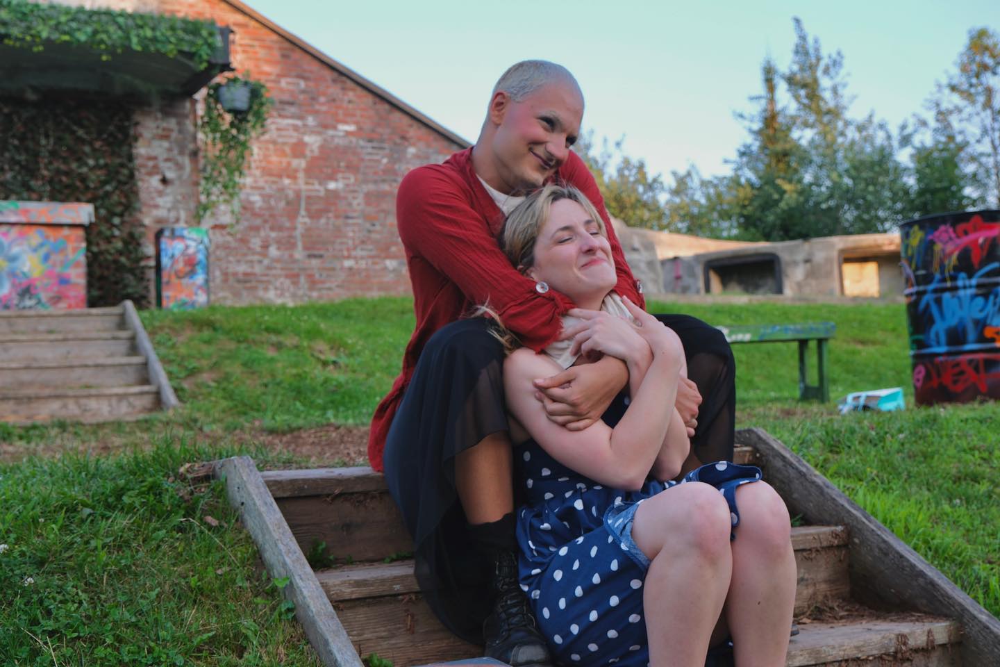 The Cambridge Battery on a beautiful clear day. In the Centre of the Image, Zach who plays The Nurse and Jade who plays Juliet sit together in a loving embrace. The Nurse is sweetly hugging Juliet. Both people look beautiful and perfectly content.