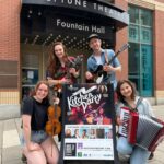 Three female musicians and one male musician crowd around a sign that says Argyle Street Kitchen Party in front of a theatre. Each musician holds a different musical instrument. They are all smiling happy smiles.