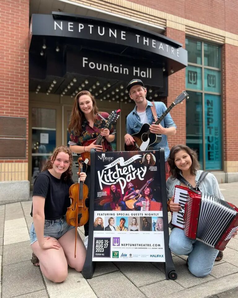 Three female musicians and one male musician crowd around a sign that says Argyle Street Kitchen Party in front of a theatre. Each musician holds a different musical instrument. They are all smiling happy smiles.