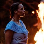 A young Black woman stands strong in front of a large campfire. She is bathed in the firelight. She wears a blue dress. She looks resolute.