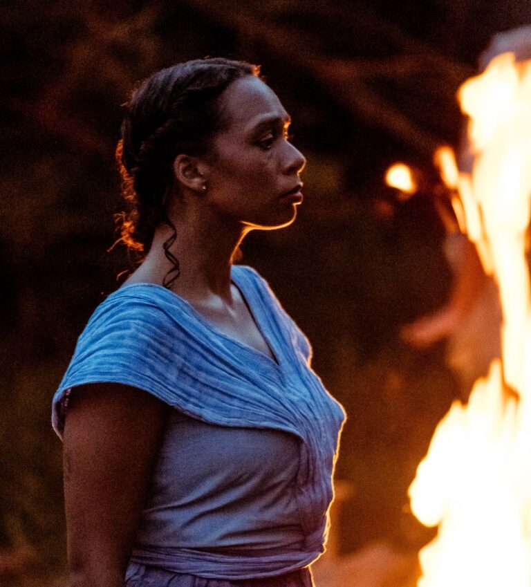 A young Black woman stands strong in front of a large campfire. She is bathed in the firelight. She wears a blue dress. She looks resolute.