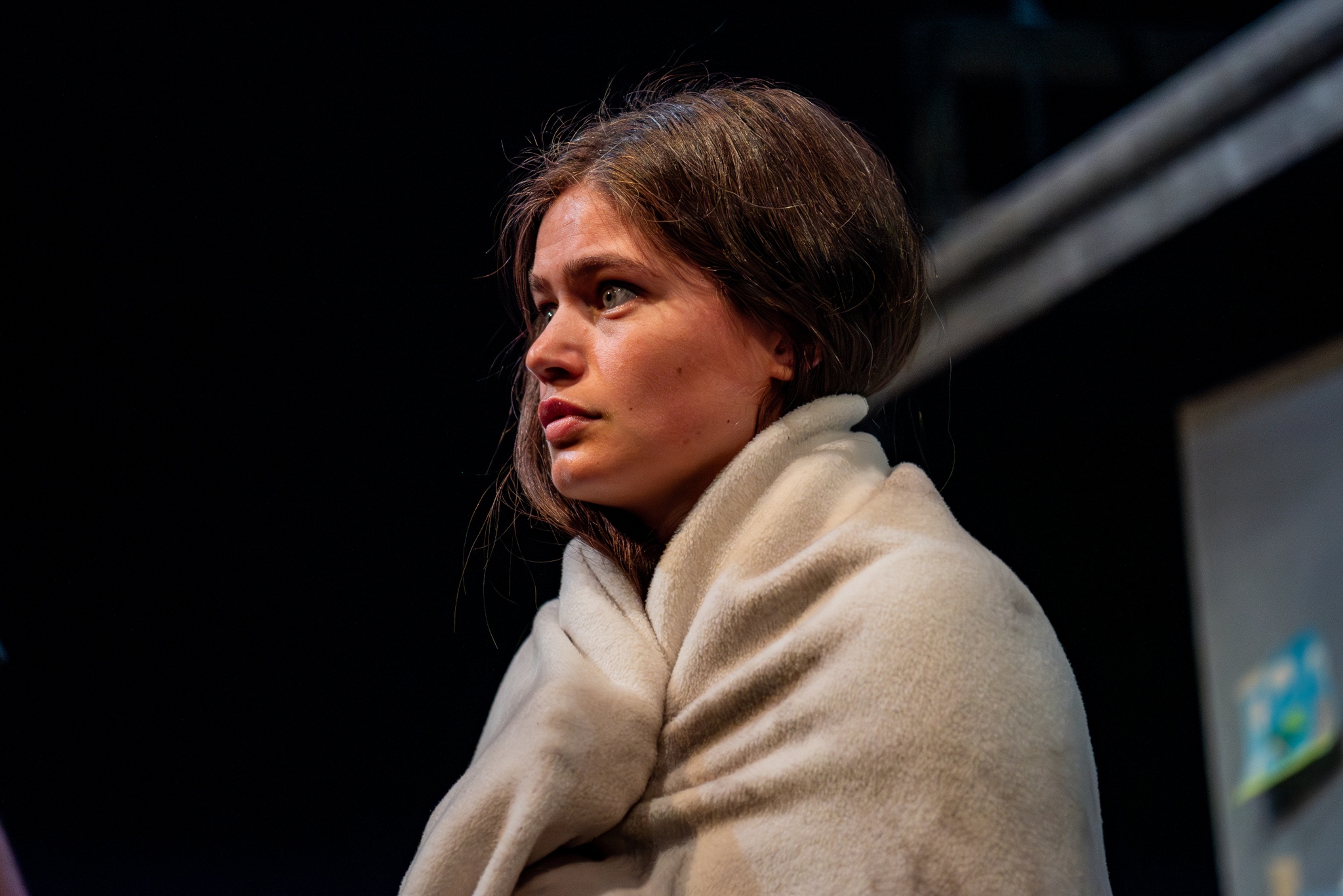 A young girl with brown hair looks very sad away from the camera, she holds an airline blanket around her.