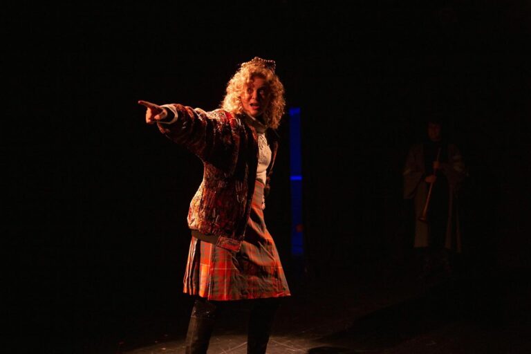 A person with red hair and wearing a crown and kilt, celebrates onstage in a golden light.