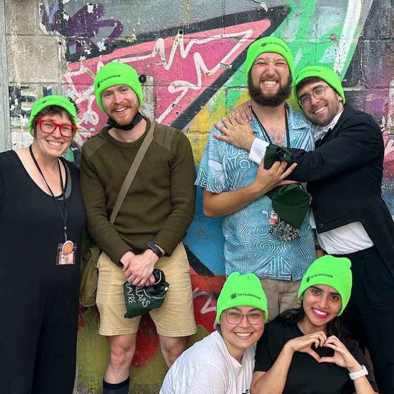Image description: a group of six people stands in front of a spray painted wall. They all wear bright green toques and smile at the camera.