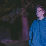 Rooks, a short white person with light brown hair and glasses, smiles while standing under a tree.