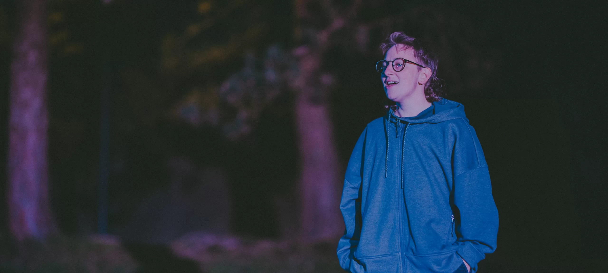 Rooks, a short white person with light brown hair and glasses, smiles while standing under a tree.