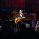A white woman, Catherine, with long hair plays the guitar and sings on a stage at a church. There are two other female musicians, Erin and Lisa, on either side of her watching her warmly.