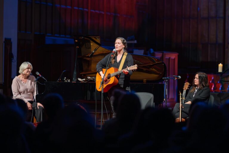 A white woman, Catherine, with long hair plays the guitar and sings on a stage at a church. There are two other female musicians, Erin and Lisa, on either side of her watching her warmly.