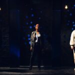 ID Three actors stand in sidelight against a black wall covered in glowing blue holes.