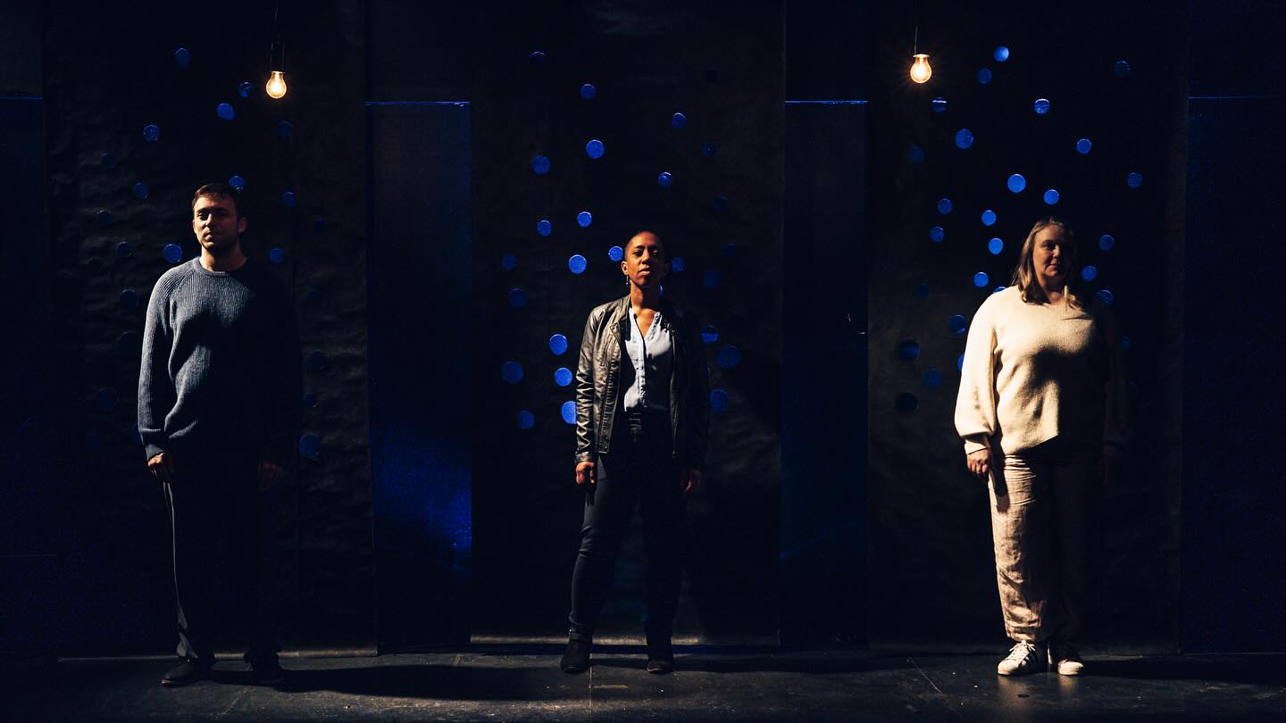 ID Three actors stand in sidelight against a black wall covered in glowing blue holes.