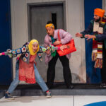 A Syrian girl in a bright yellow hijab slipping on ice in a curling rink. A Black woman and Indian man in bright warm clothes rush to help her so she doesn't fall.