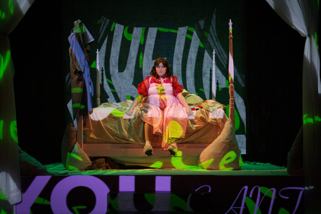 Juliet in bright red and pale pink, and with dark hair looking a bit like Little Red Riding Hood in a large pink bed, surrounded by projections in green, blue, and purple that are words we can’t quite make out.