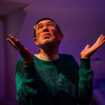 A Queer man in a green sweater and glasses looks up with a serious expression on his face. Both his elbows are bent and his hands are extended upward and outward, almost a shrug, except his shoulders are not raised.