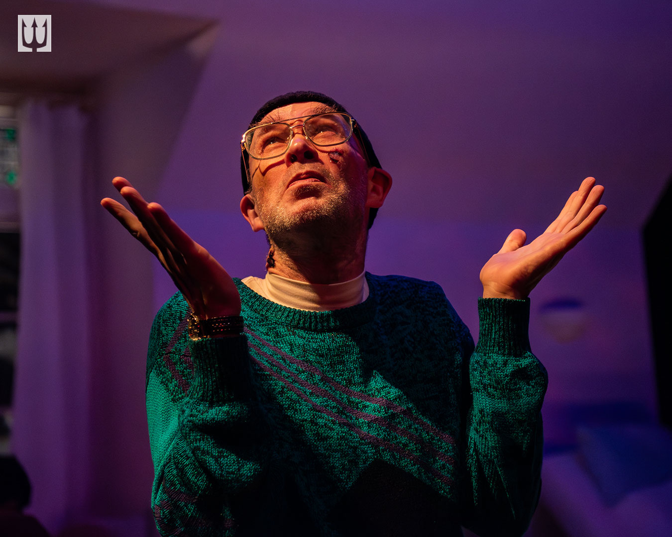 A Queer man in a green sweater and glasses looks up with a serious expression on his face. Both his elbows are bent and his hands are extended upward and outward, almost a shrug, except his shoulders are not raised.