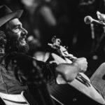 Adam Baldwin, a white man in his late 30s with a dark beard grins while playing the guitar and wearing a cowboy type hat. He is facing upstage from the camera, but you can still see his happiness clearly.