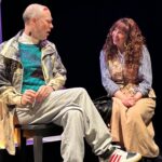 A gay man dying from AIDS, he has lesions on his face and wears baggy clothes, he sits facing his sister, a healthier looking woman in her 30s with brown, long, curly hair.