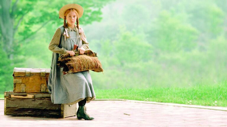 Megan Follows as 12 year old Anne Shirley, a thin orphan with red hair in braids, sitting at a Victorian train station with her carpet bag.