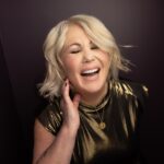 Jann Arden, a white woman with short blond hair laughs with her eyes closed and her right hand held up close to her right cheek. She is wearing a fancy gold top. She looks genuinely happy.