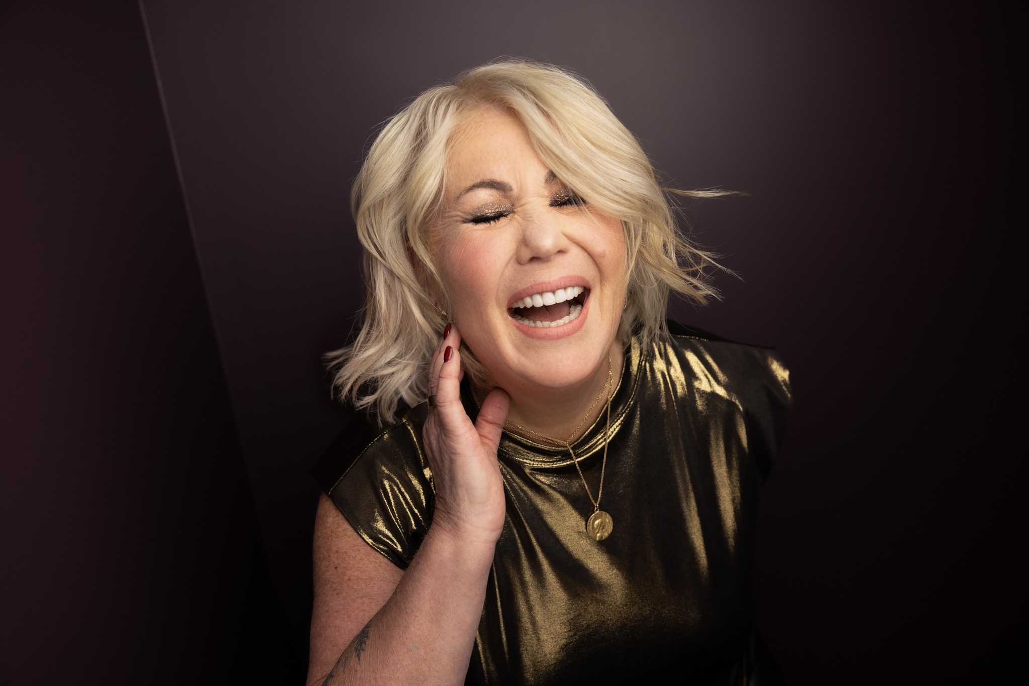 Jann Arden, a white woman with short blond hair laughs with her eyes closed and her right hand held up close to her right cheek. She is wearing a fancy gold top. She looks genuinely happy.