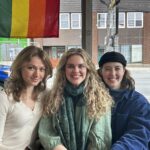 A cute photo of three beautiful young friends in front of a window with a Pride flag hanging behind them. Ella and Riley have long light hair, Jessie has long brown hair. Riley is wearing a big scarf. Jessie is wearing a beret. Ella looks enticingly at the camera while Riley and Jessie grin.