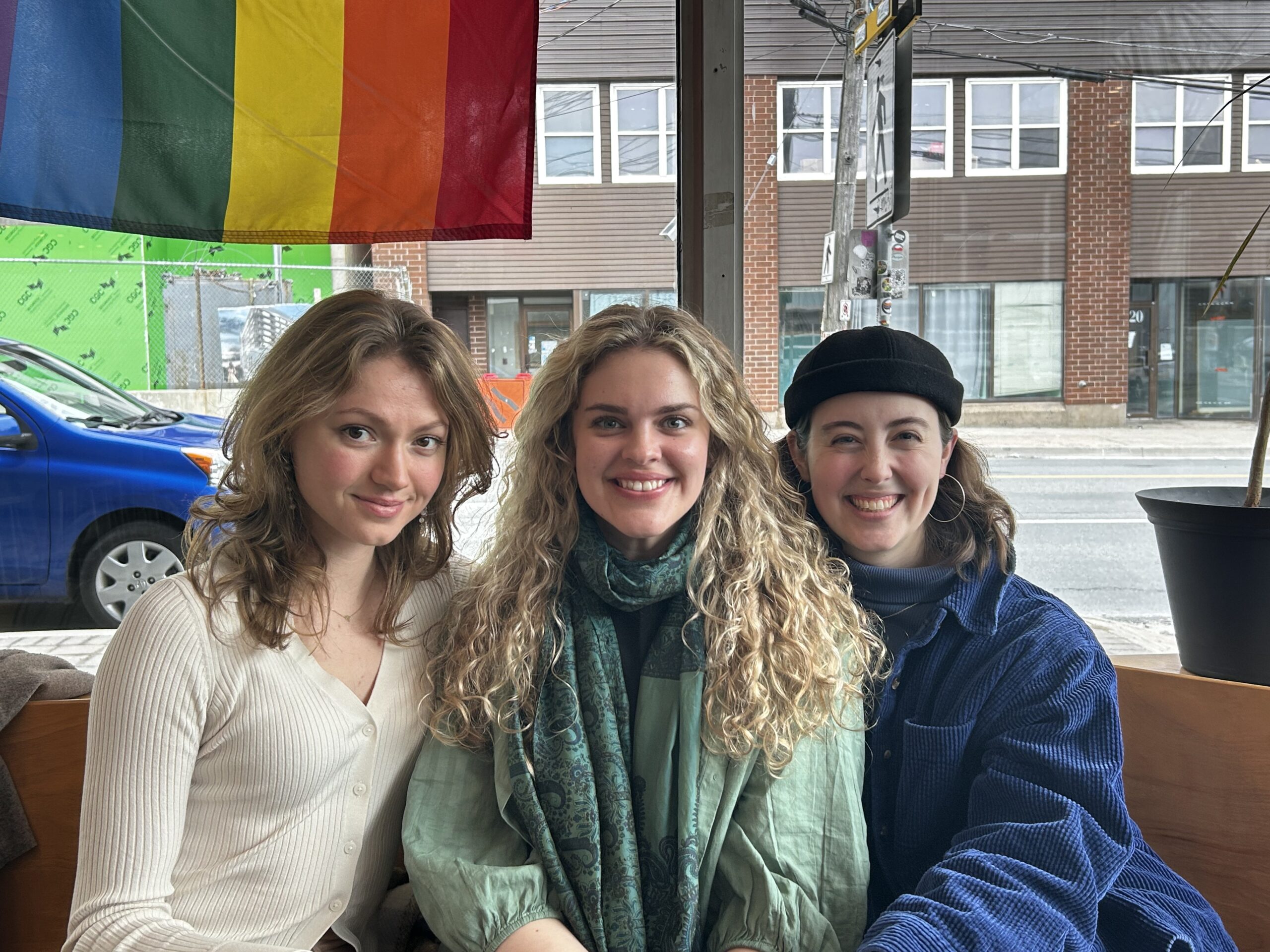 A cute photo of three beautiful young friends in front of a window with a Pride flag hanging behind them. Ella and Riley have long light hair, Jessie has long brown hair. Riley is wearing a big scarf. Jessie is wearing a beret. Ella looks enticingly at the camera while Riley and Jessie grin.