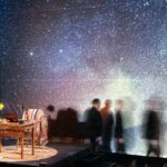 An extremely starry background. A small desk with a green lamp on it and a chair with a colourful knit blanket looks small to the left. To the right various images- almost shadows becoming more faded as they move to the right- of the protagonist Fred. We just see the outline of him.