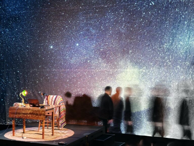 An extremely starry background. A small desk with a green lamp on it and a chair with a colourful knit blanket looks small to the left. To the right various images- almost shadows becoming more faded as they move to the right- of the protagonist Fred. We just see the outline of him.