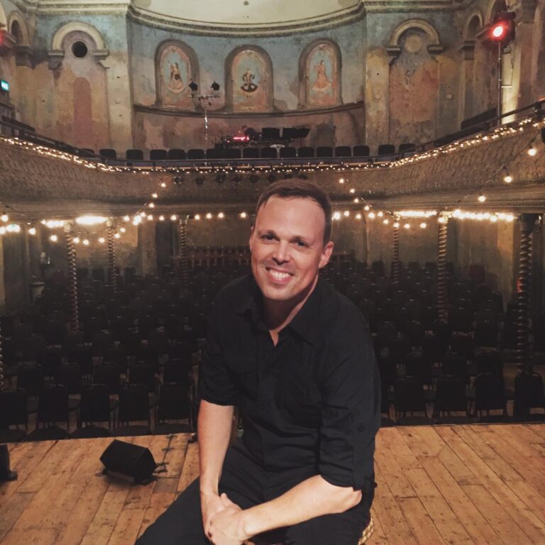 A smiling man with a moustache and short light hair is dressed all in black and sits on a stage in an ornate theatre