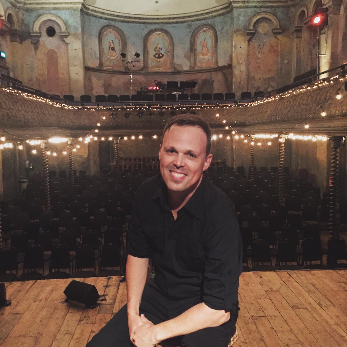 A smiling man with a moustache and short light hair is dressed all in black and sits on a stage in an ornate theatre