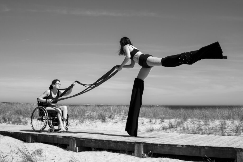 pril and Vanessa battle for power during a circus performance on the boardwalk at a local beach. April sits in her wheelchair while Vanessa towers over her on stilts.